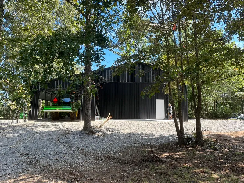 Black vertical-roof barn set among trees on a gravel pad