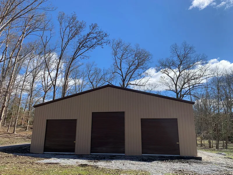 Tan vertical-roof garage with three brown roll-up doors under a blue sky